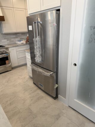 Stainless steel French-door refrigerator with protective plastic wrap installed in a Frisco kitchen remodel with white cabinetry and tile flooring.